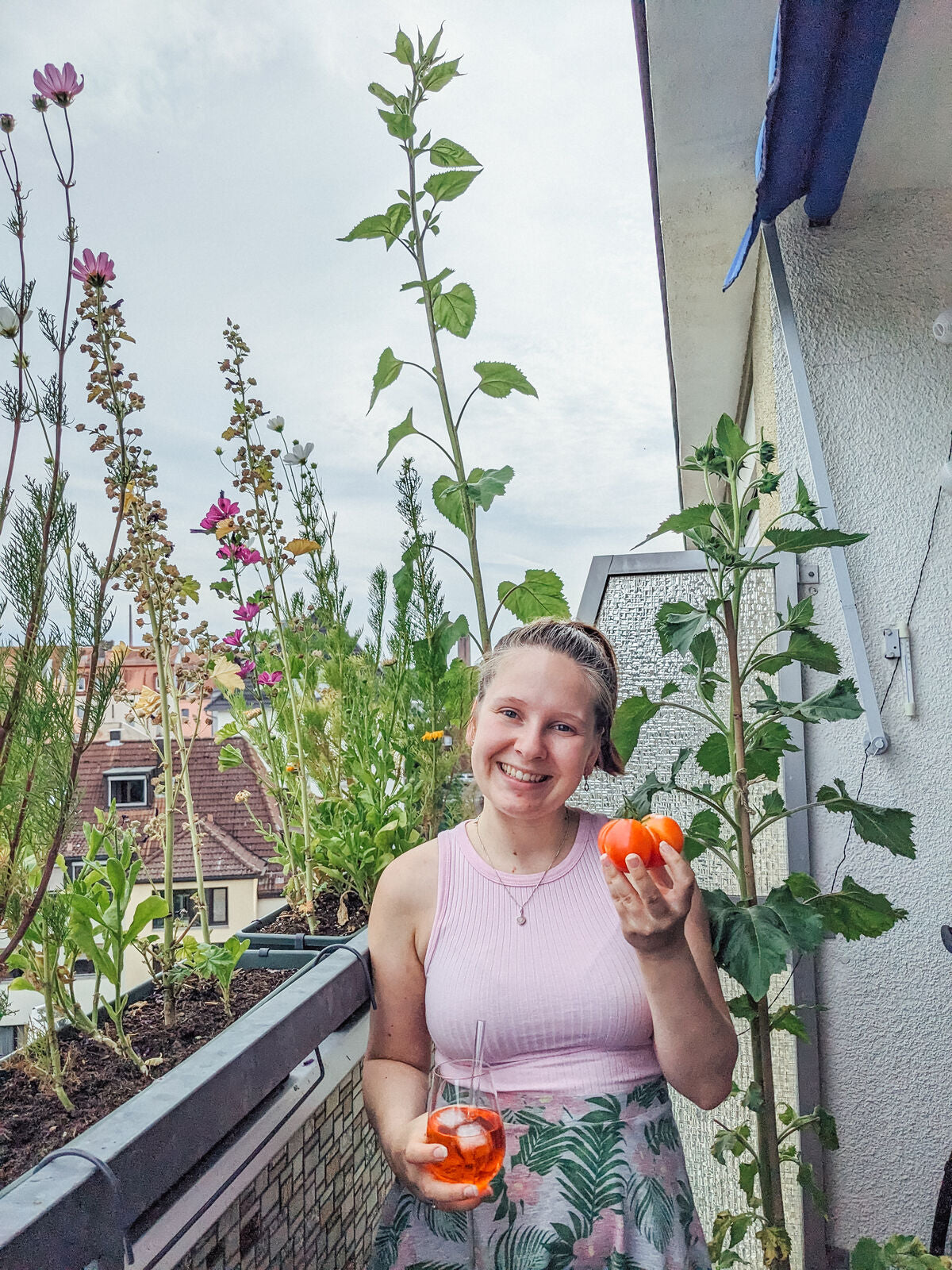 Masha hält eine Tomate und steht vor vielen Pflanzen (u.a. in Blumenkästen)